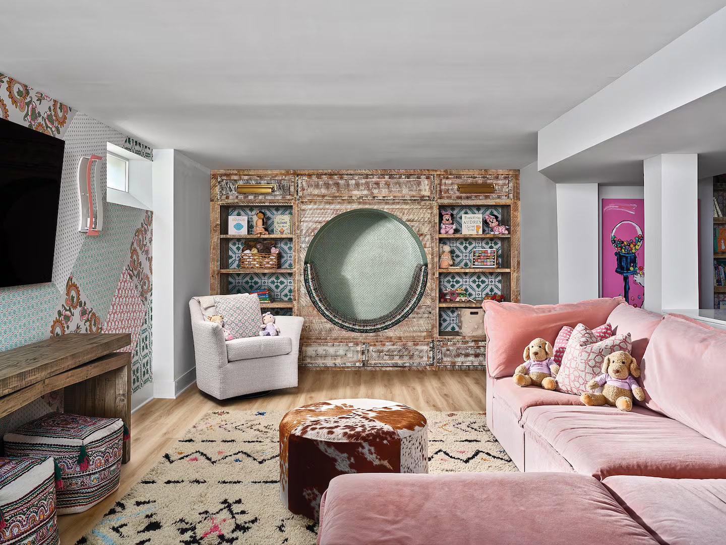 A room with built-in, circular book nook in the center of the back wall. On either side of the nook are built-in bookshelves. In front of the bookshelf on the left, there's a cozy chair. On the right side of the room, there's a pink sectional sofa. In front of that there's a patterned rug and an ottoman that looks like cowhide. On the left wall, there's a low table, a wall-mounted TV, and a small basement window toward the top of the wall.